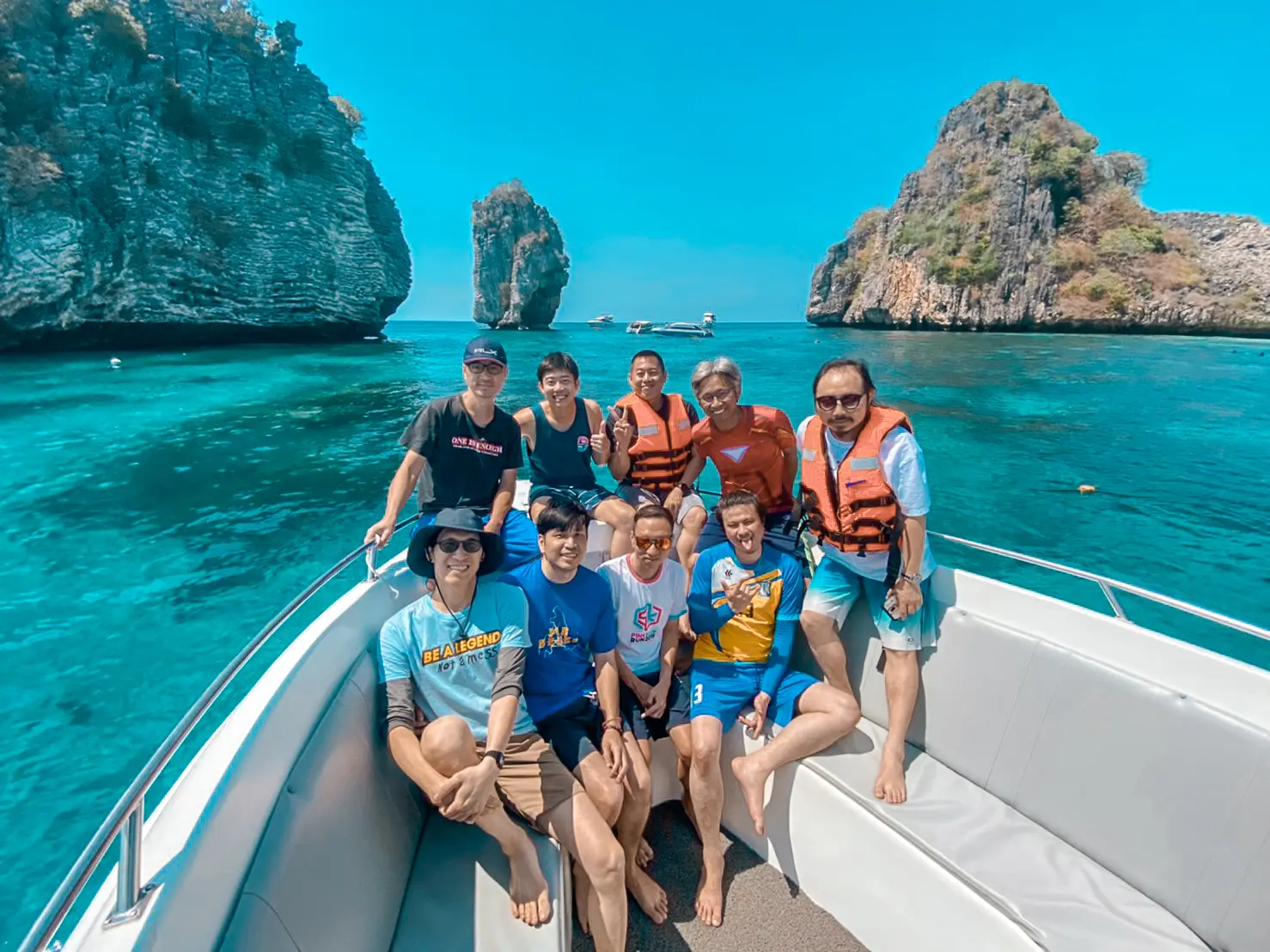 Men in T-shirts and orange life vests crowding a speedboat's bow over turquoise water near limestone islets