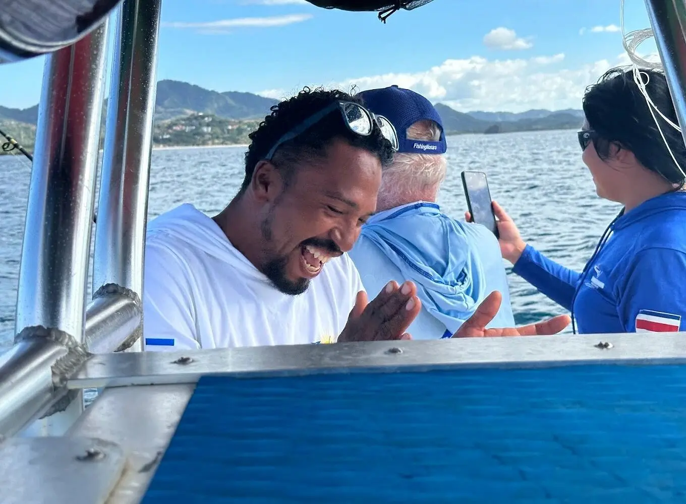 Guests laughing together on the fishing boat as one checks a phone, green hills behind
