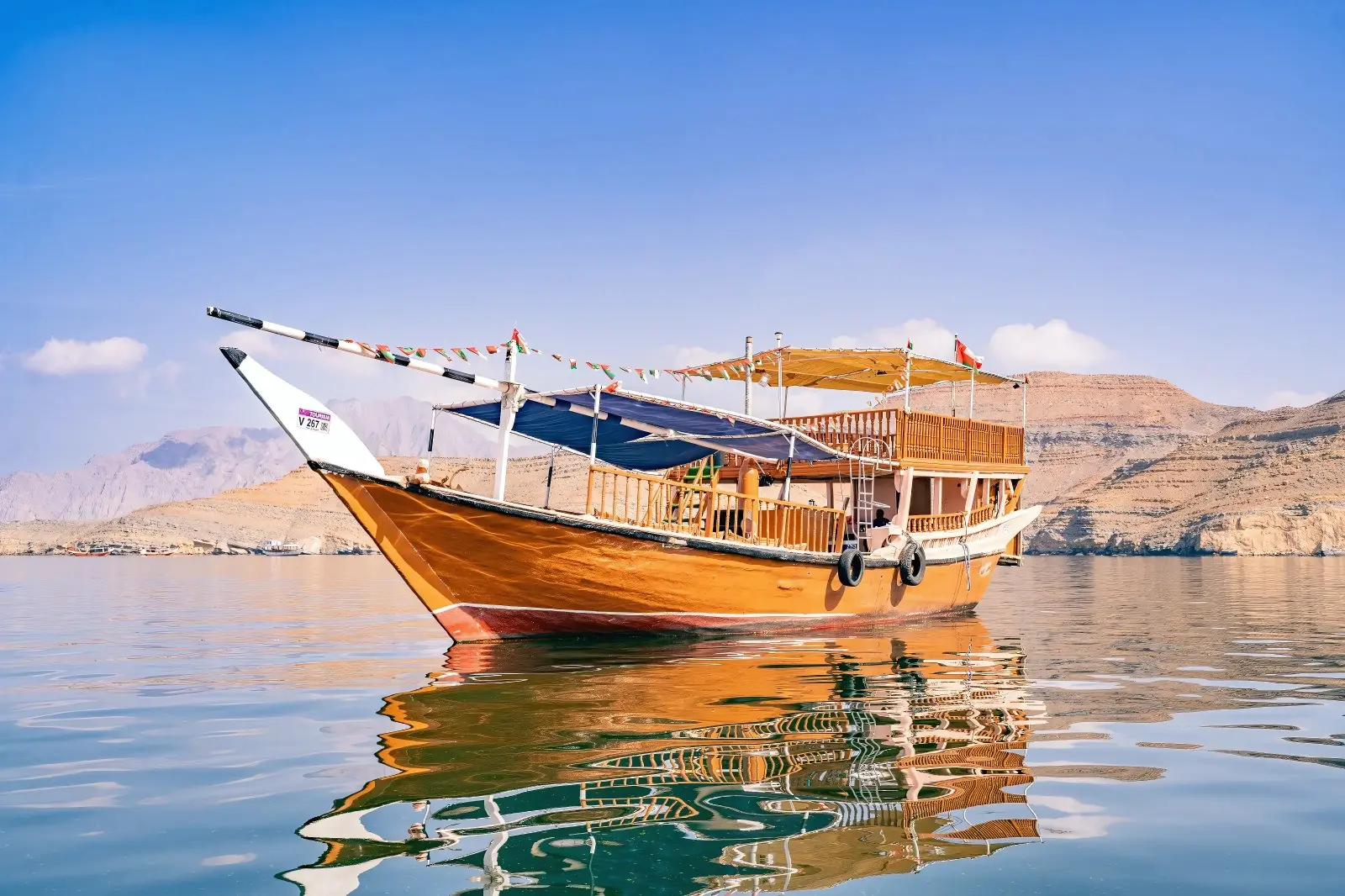 Wooden dhow anchored on glassy water, its hull reflected below the bare Musandam mountains