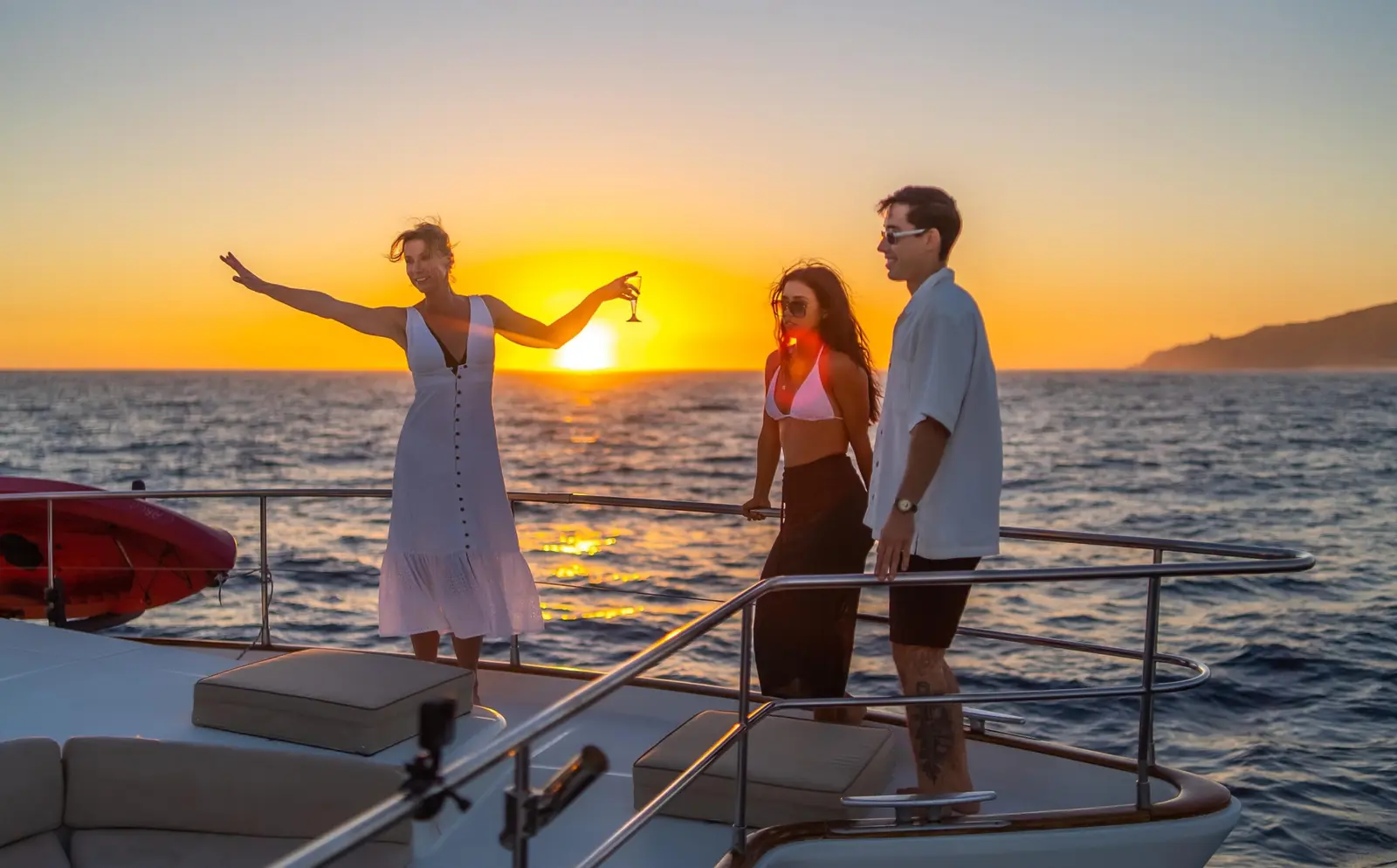Woman in a white dress dancing on a catamaran's bow at sunset off Cabo San Lucas as a couple looks on