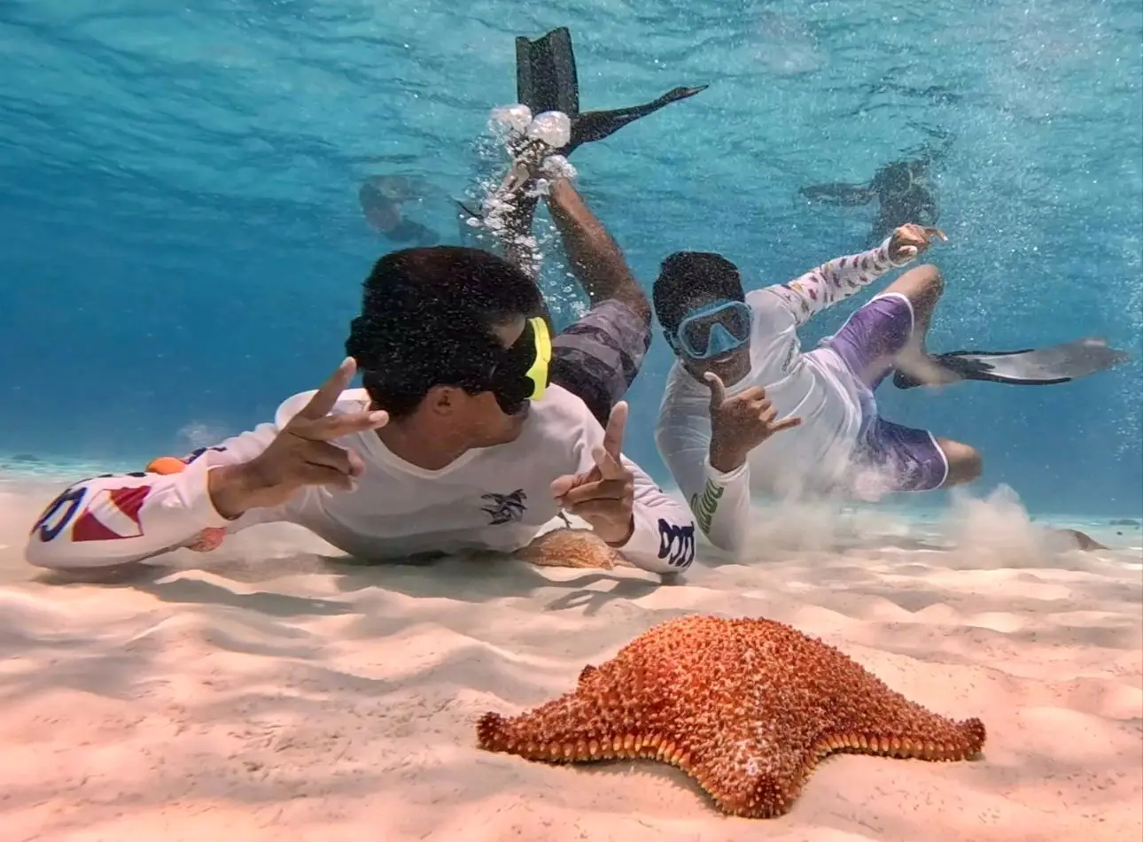 Two snorkelers flash hand signs on the sandy seabed next to a large orange starfish