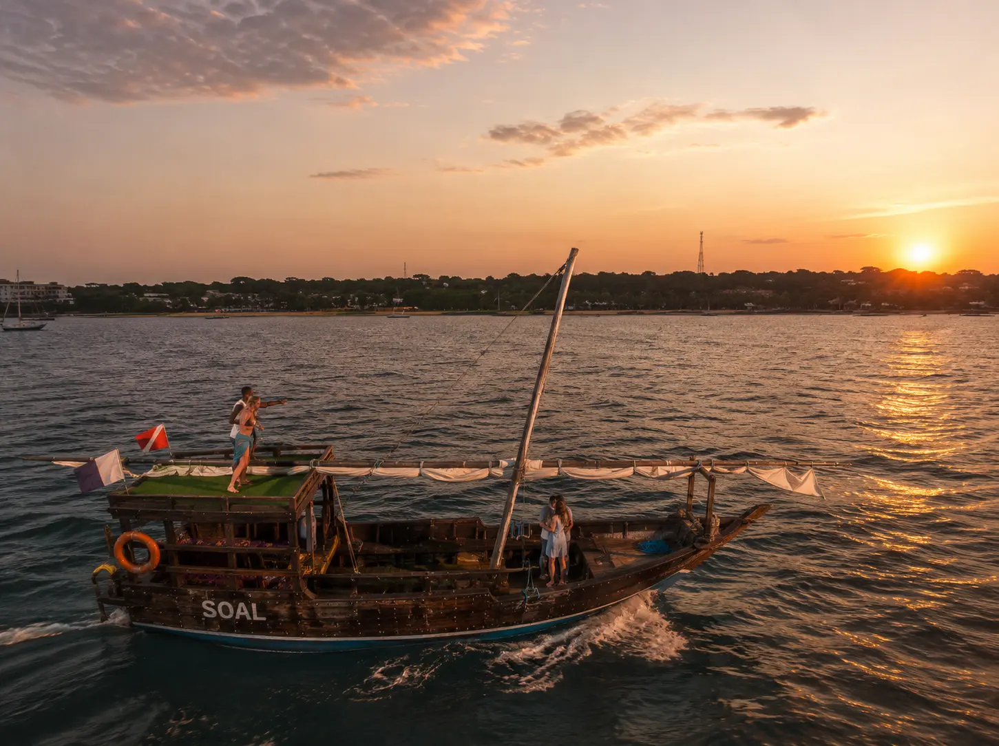 Two-deck dhow with its sail furled cruising toward Nungwi at sunset, a figure on the rooftop deck