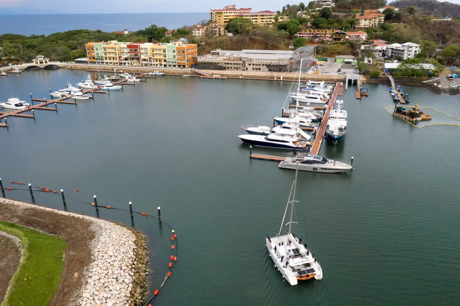 Aerial view of a sailing catamaran in the Playa Flamingo marina past docked yachts and a rock breakwater
