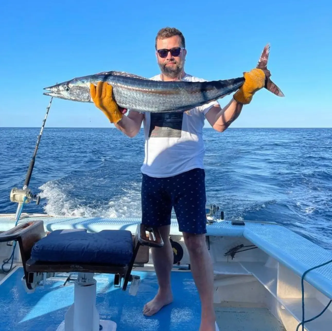 A man in gloves holds up a long silver fish on the back deck of the fishing boat