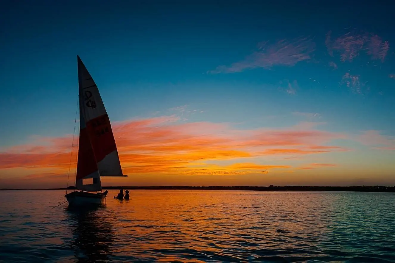 Small sailboat with a red-and-white sail on calm Bacalar Lagoon under a glowing orange sky