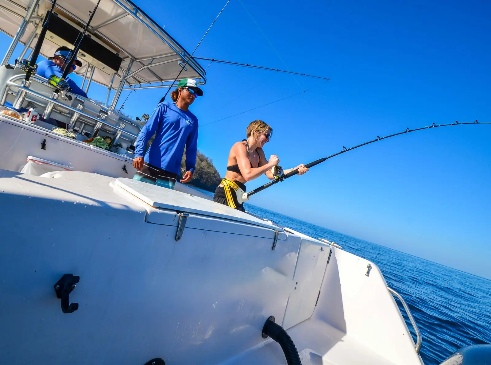 A woman fights a fish on a bent rod beside a man in a blue shirt on the deck of the fishing boat