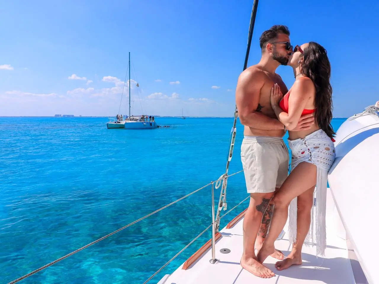 Couple kissing on a sailing catamaran deck with another catamaran anchored in turquoise water