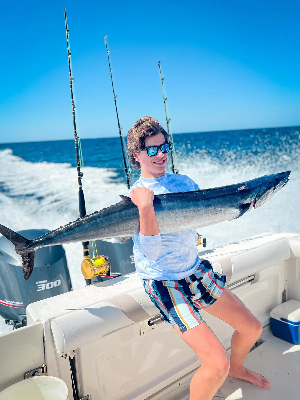 Angler in sunglasses holding up a long wahoo at the stern of the center console boat as it runs offshore