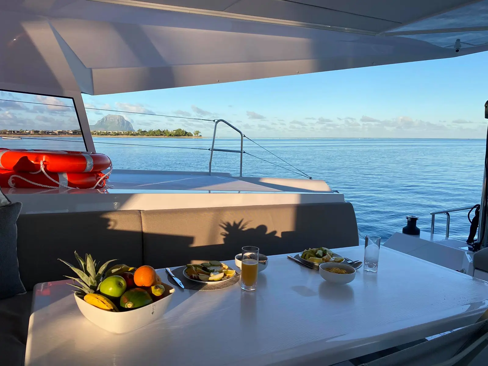 Fruit bowl, drinks and snacks laid out on a yacht's shaded deck table overlooking the sea