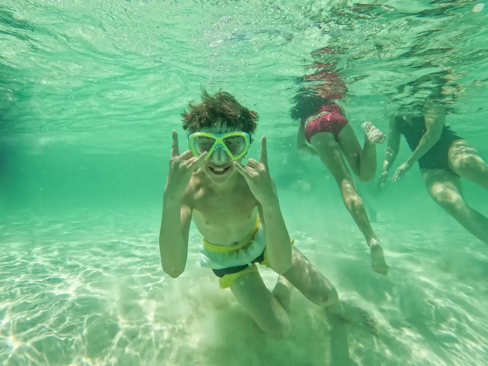 Boy in a snorkel mask making hand signs underwater as others swim behind