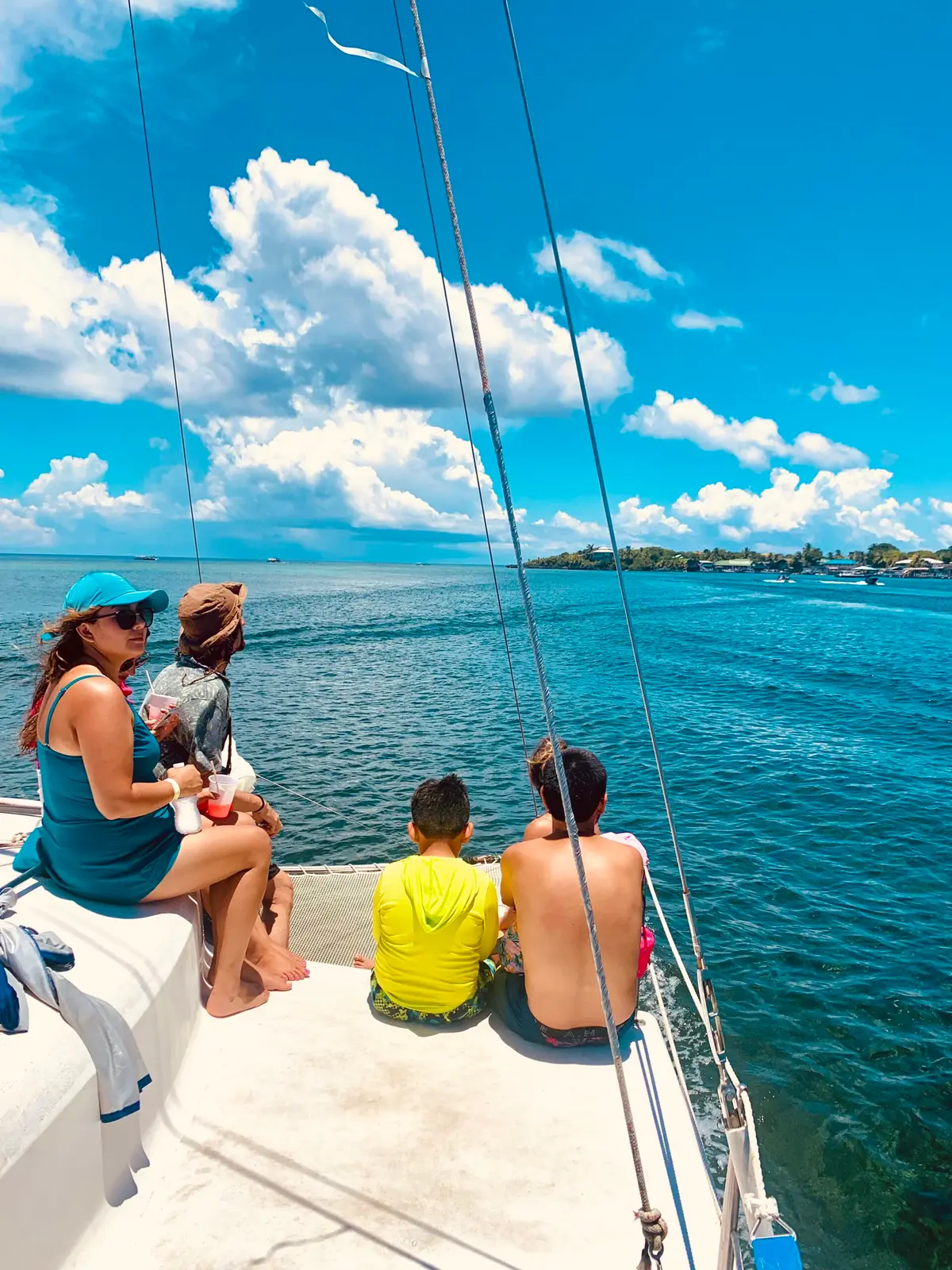 Guests seen from behind sitting on the catamaran's bow, gazing over blue water toward Roatan
