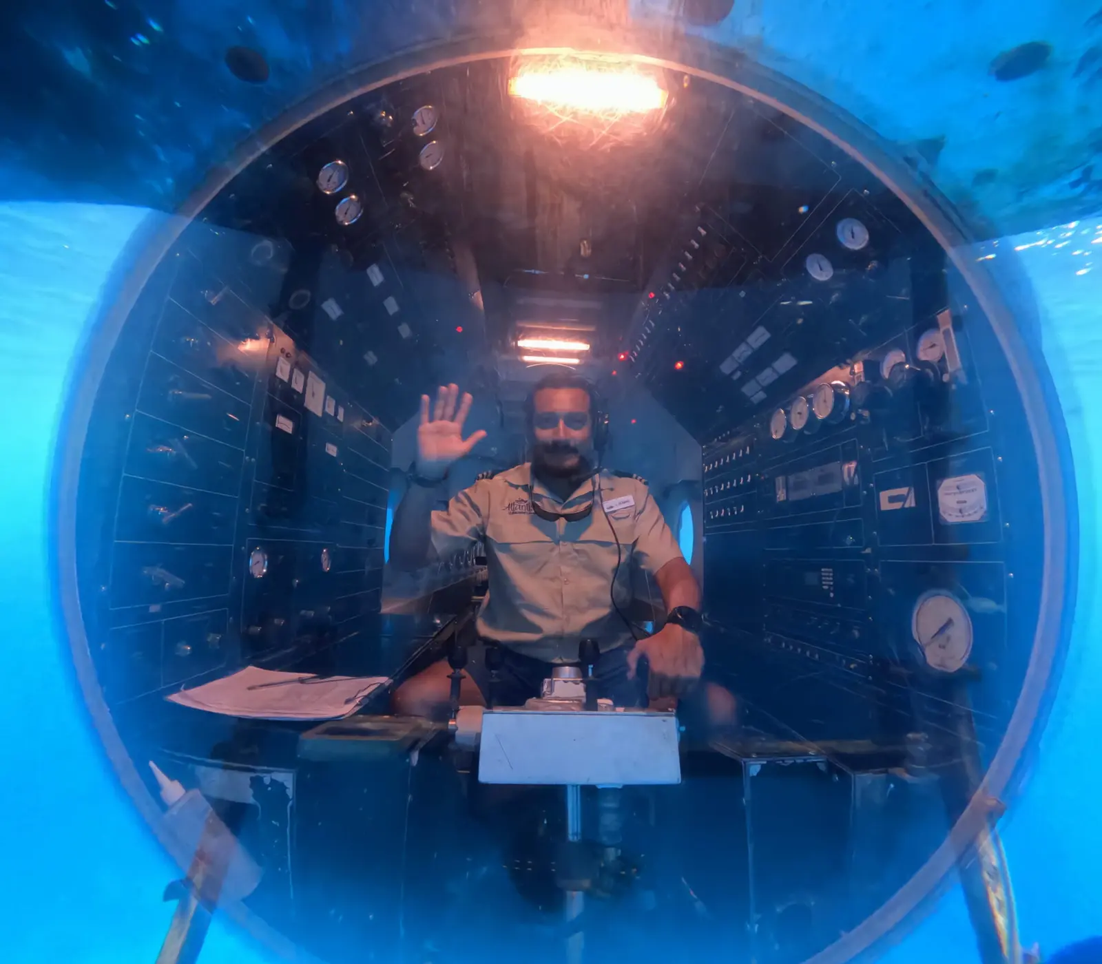 Submarine pilot waving through the round front viewport, seen from the water outside