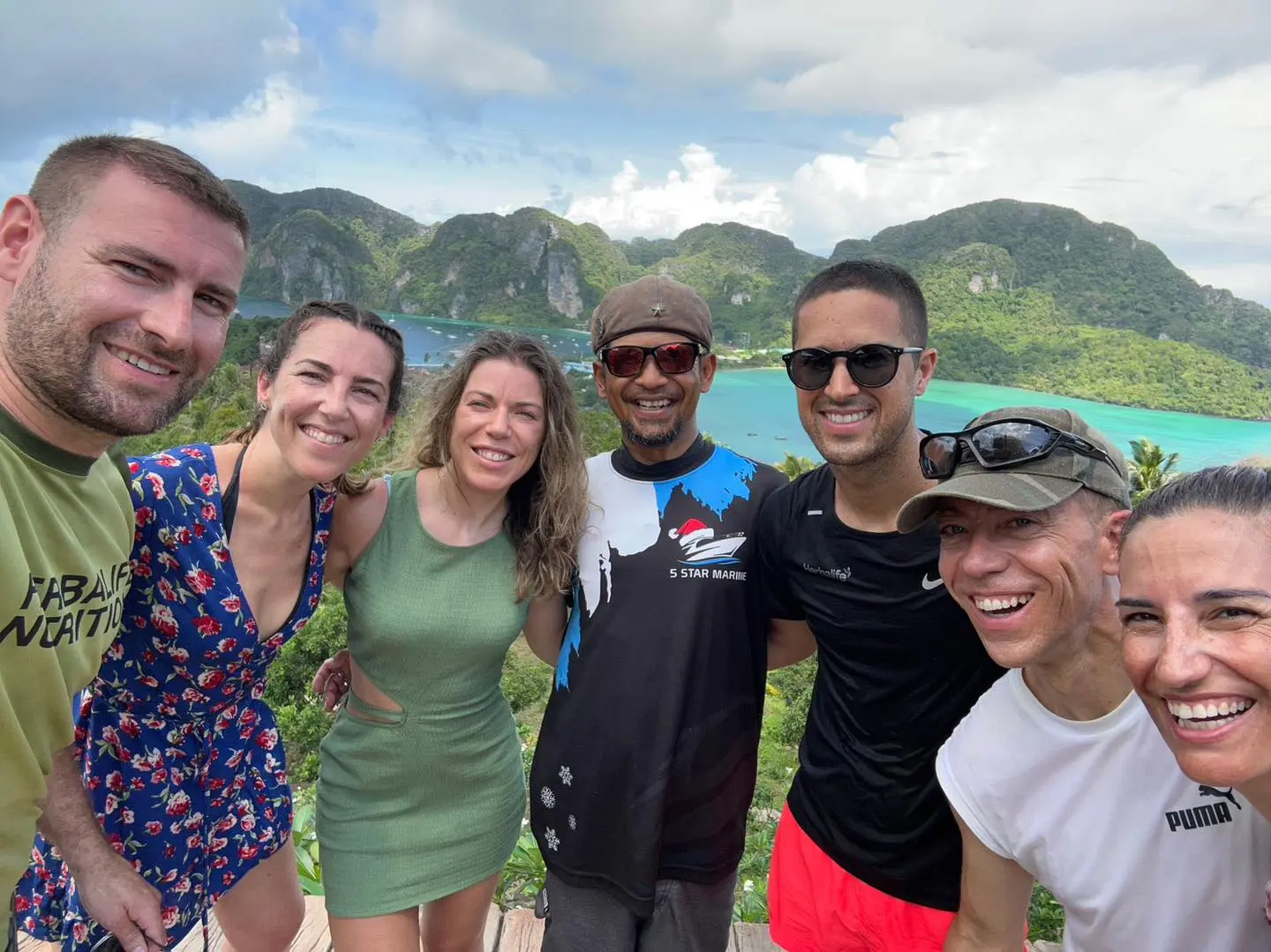 Friends taking a group selfie at a hilltop viewpoint above green hills and turquoise water in Thailand