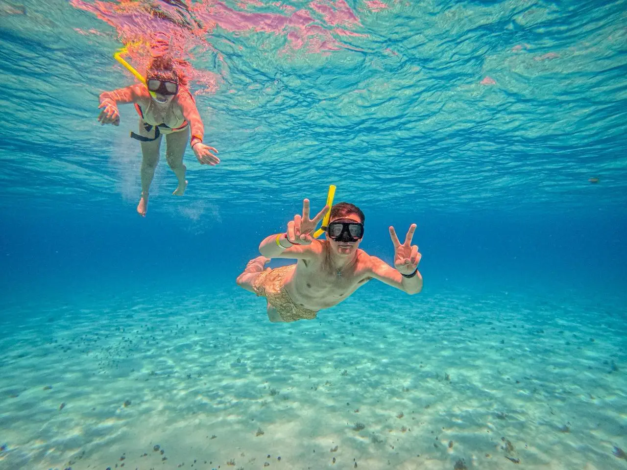 Two snorkelers underwater, one flashing peace signs above the white sandy seabed