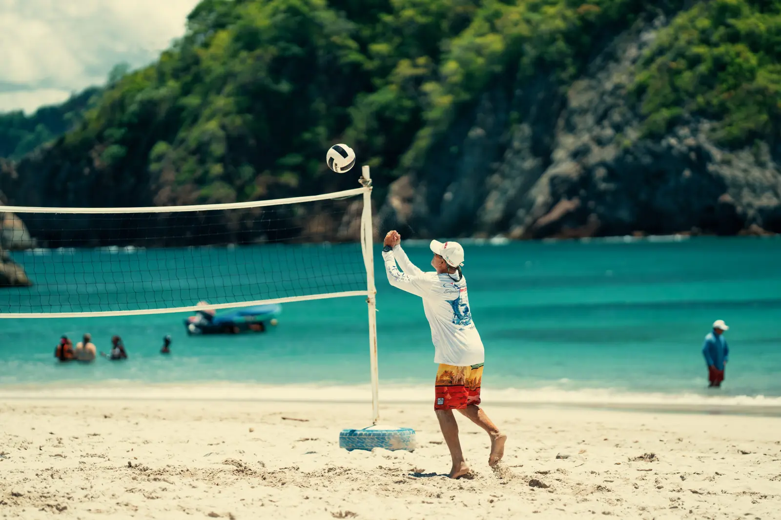 Man hitting a volleyball over the net on Tortuga Island's white sand, swimmers in the turquoise bay