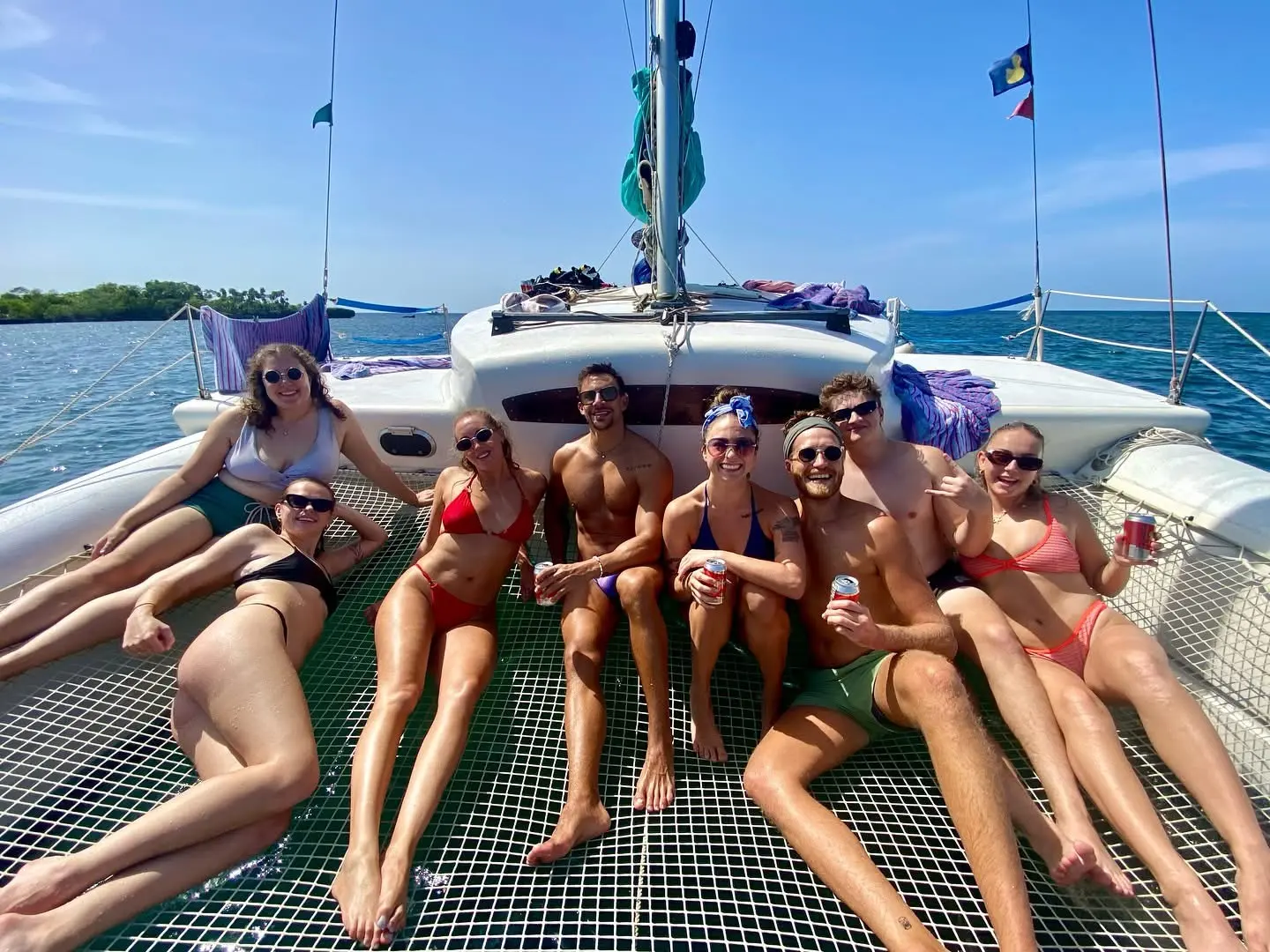 Friends in swimwear lounging on the catamaran's trampoline net beneath the mast, Roatan shore behind