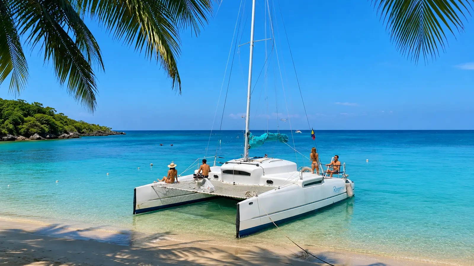 Guests relaxing aboard a sailing catamaran anchored in turquoise water off a palm-shaded Roatan beach