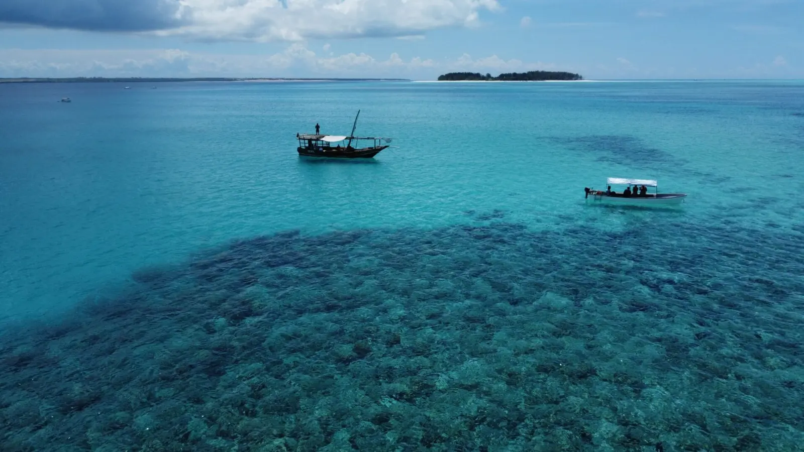 Dhow and a small motorboat anchored above a dark coral reef in clear turquoise water off Zanzibar