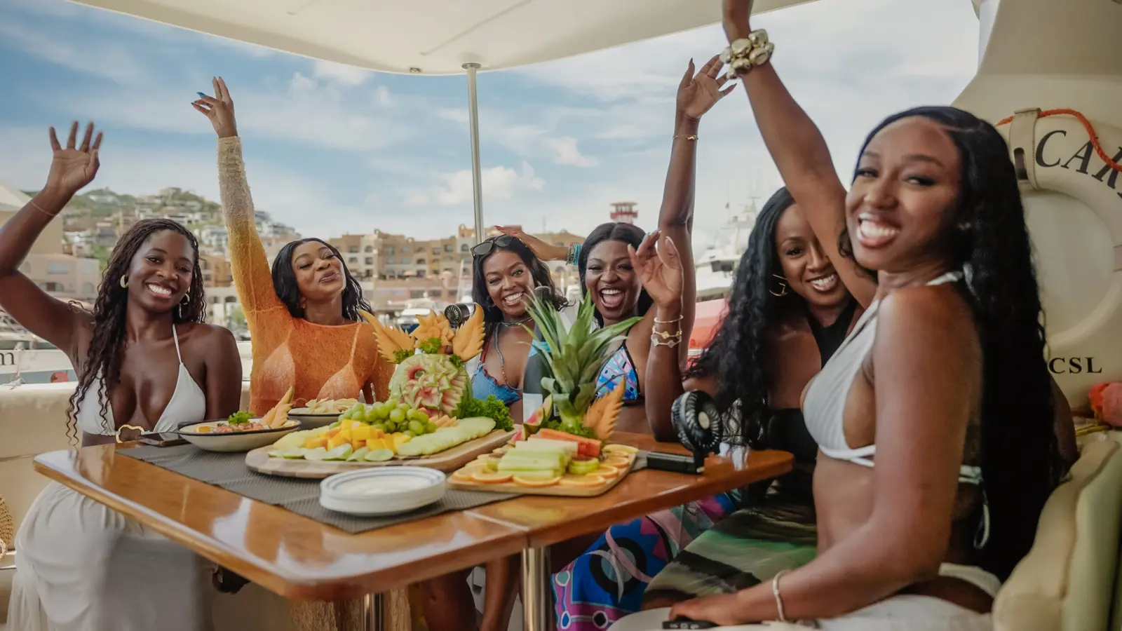 Women cheer around a table spread with fruit platters and snacks aboard the catamaran
