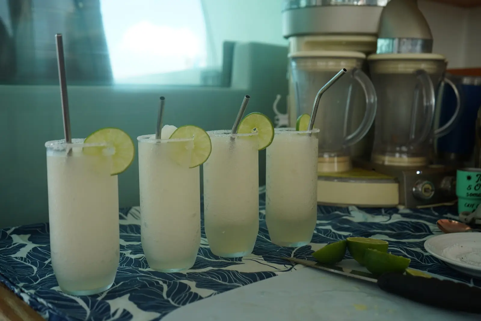 Four frozen margaritas with lime wedges and steel straws beside blenders in the galley
