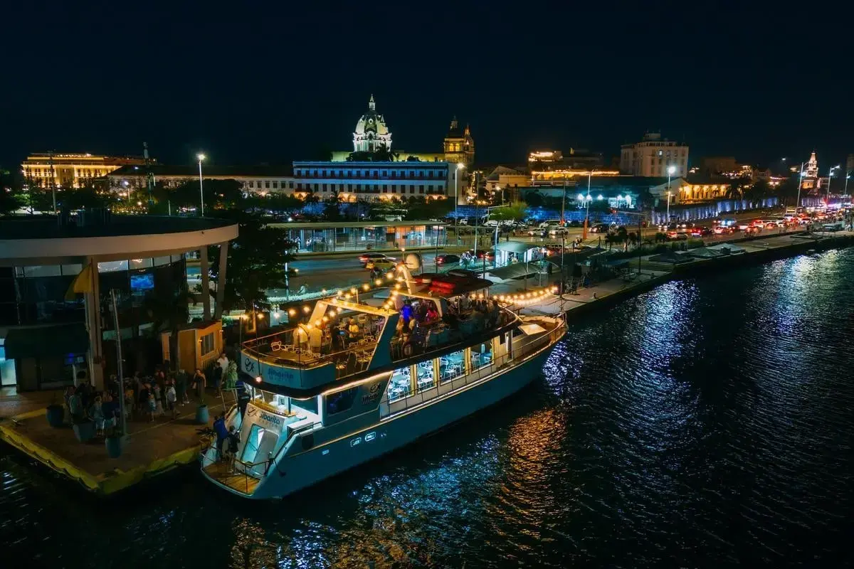 Two-deck motor yacht lit up at night beside a pier in Cartagena, the walled city's dome glowing behind