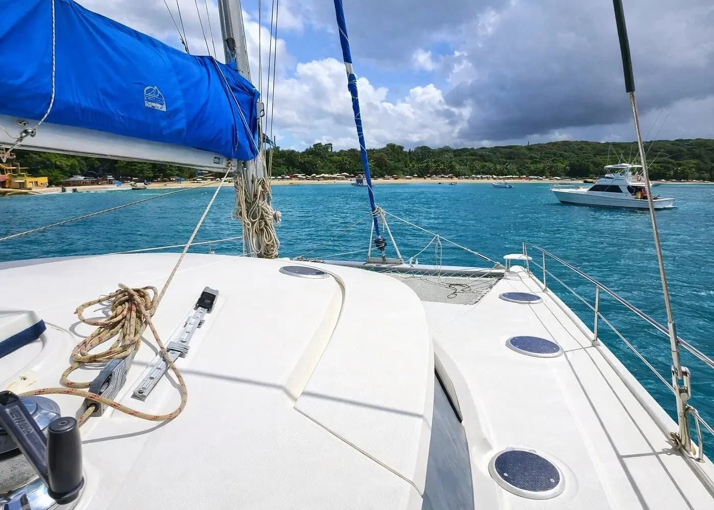 Foredeck of a sailing catamaran with a blue sail cover, anchored off the green Puerto Plata coast