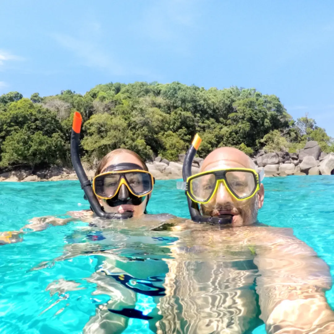 Two snorkelers in yellow masks taking a selfie in clear water near a rocky islet