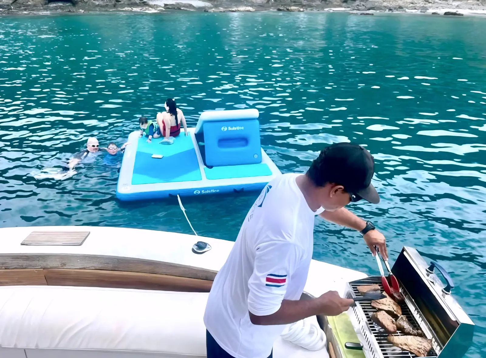 Crew member grilling at the stern as guests lounge on an inflatable floating platform in a Guanacaste cove