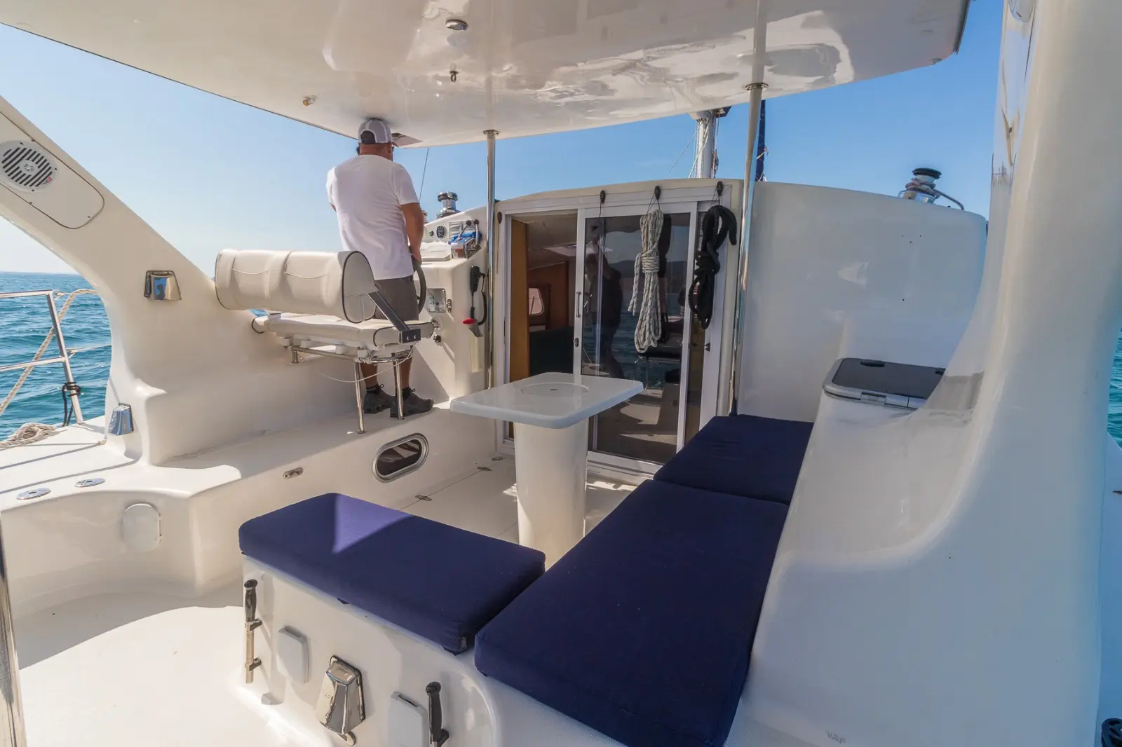 Shaded aft deck of a catamaran with navy cushions and a table as the captain stands at the helm