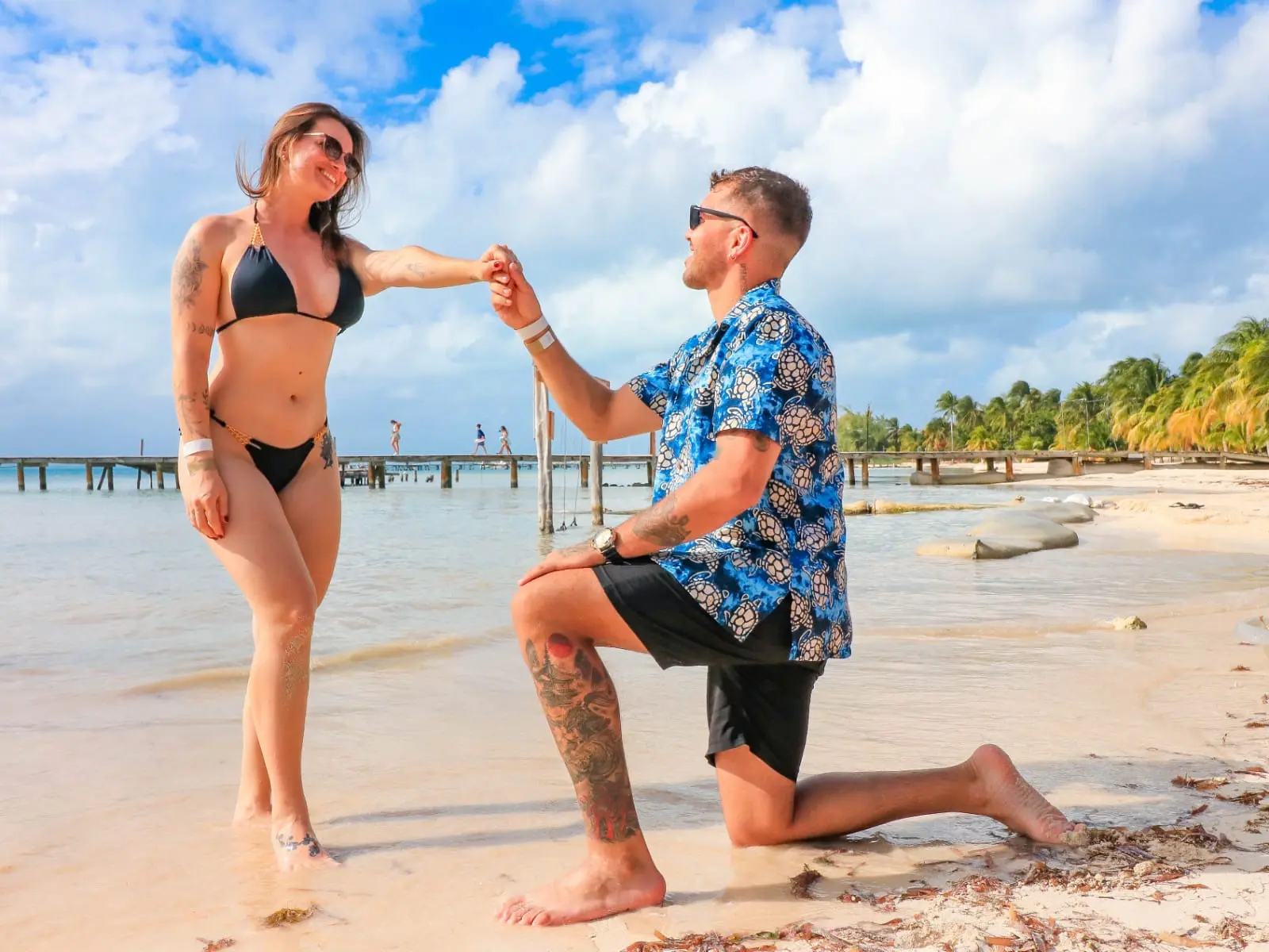 Man on one knee proposing to a woman in a black bikini on an Isla Mujeres beach by a wooden pier