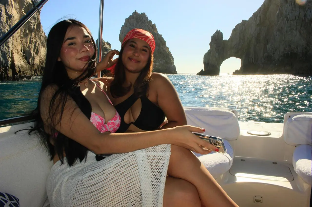 Two women in swimsuits pose together on the yacht's cushioned seat with El Arco behind them