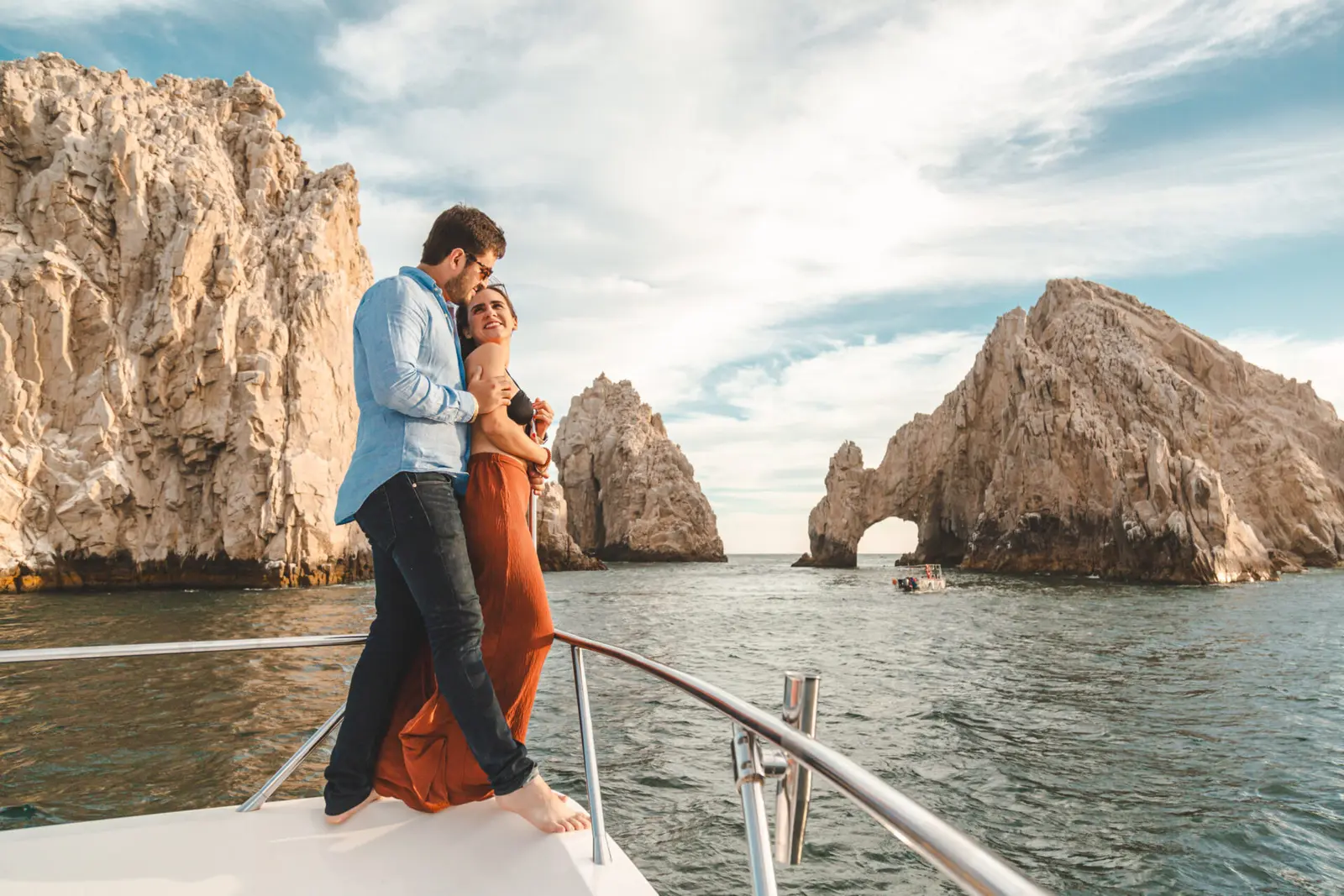 A couple embraces at the yacht's bow rail with the arch and granite cliffs of Los Cabos behind
