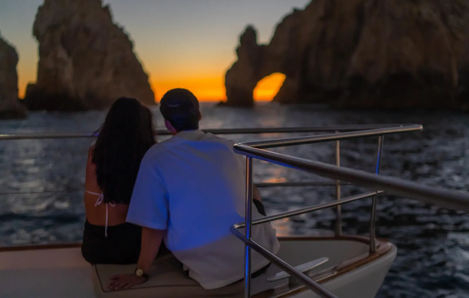 Couple sitting at a catamaran's bow rail watching the sun set through El Arco