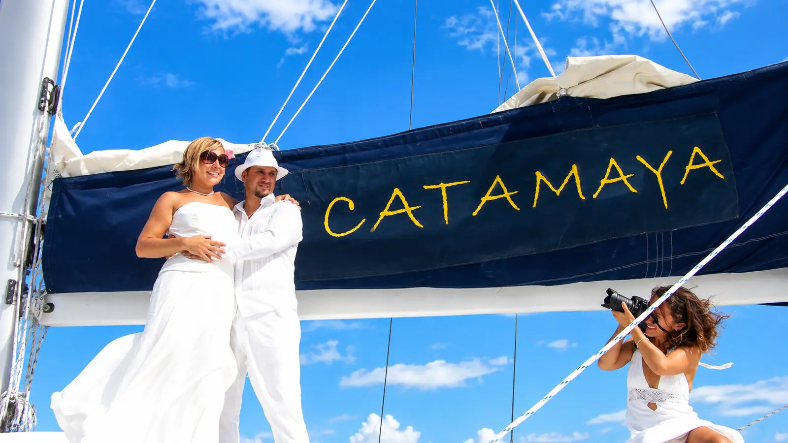 Bride and groom in white posing by the mast of a sailing catamaran as a photographer kneels to shoot them