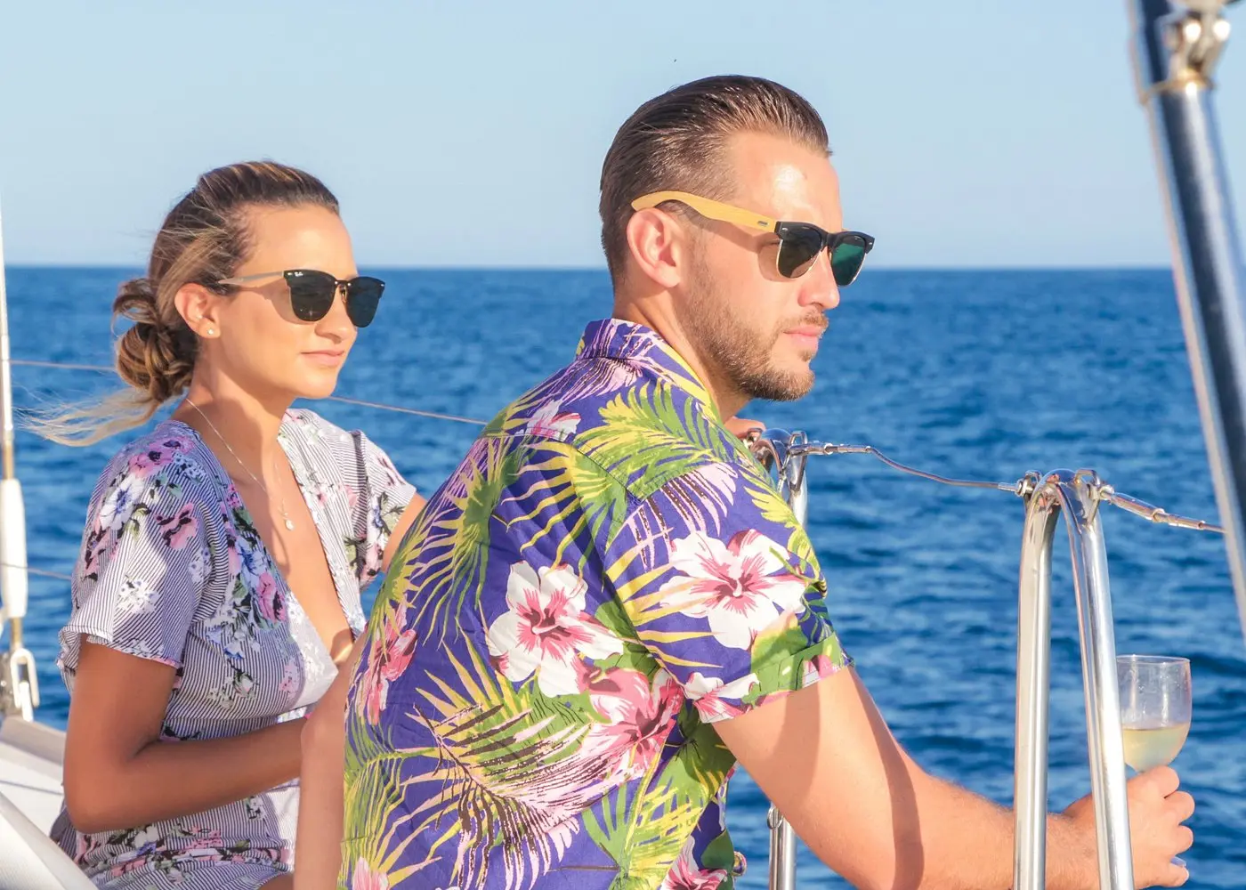 Couple in floral shirts and sunglasses at the rail of a catamaran, one holding a glass of wine