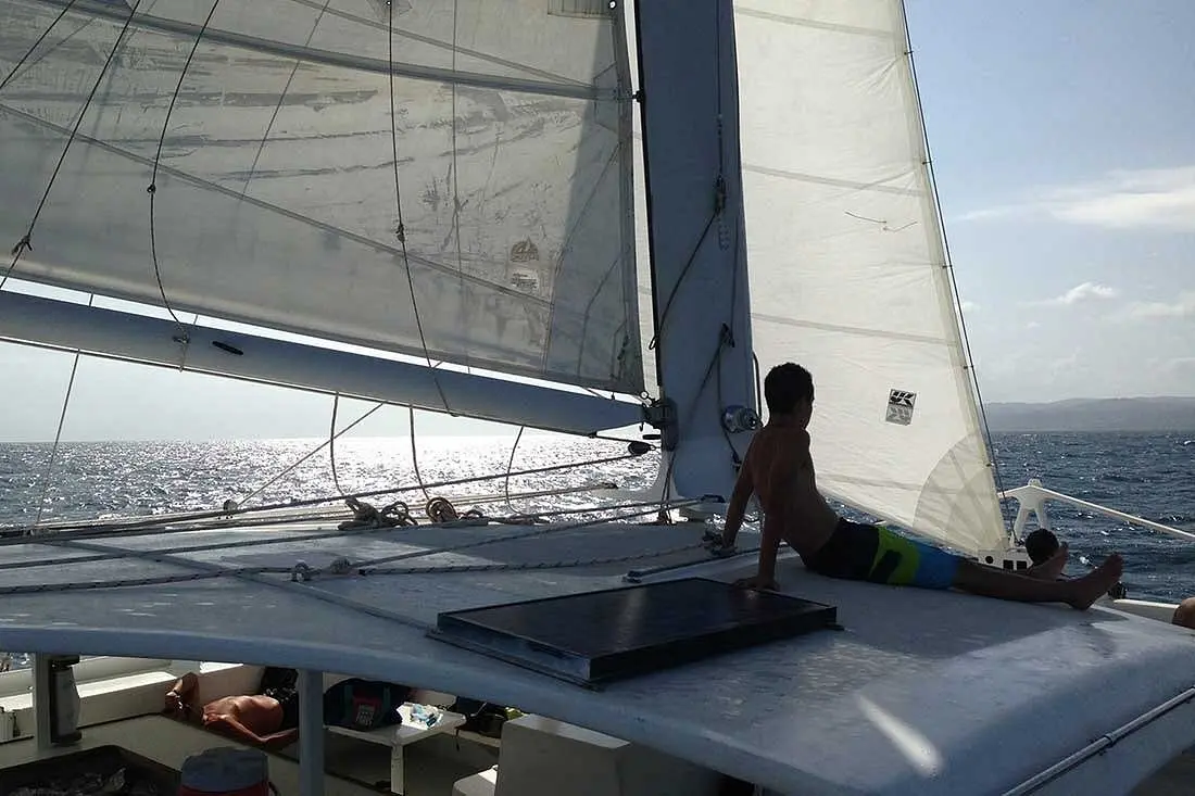 Boy sitting on the catamaran's cabin roof beneath the raised sails, sunlit sea beyond
