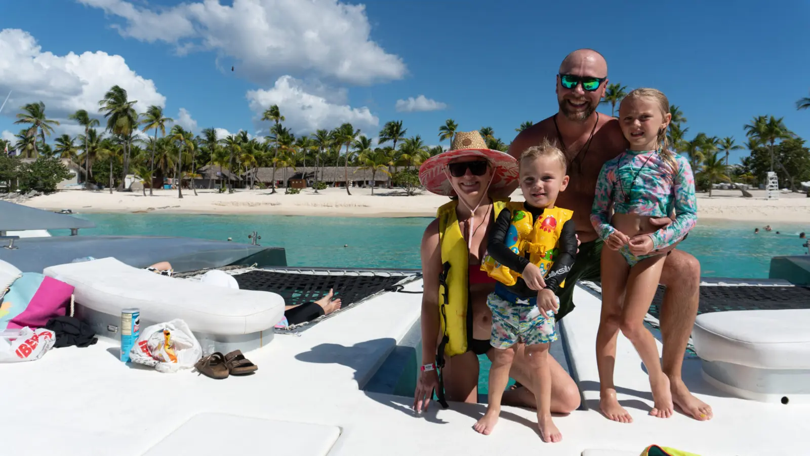 Family with two kids posing on the catamaran's front deck, a palm-lined beach behind them