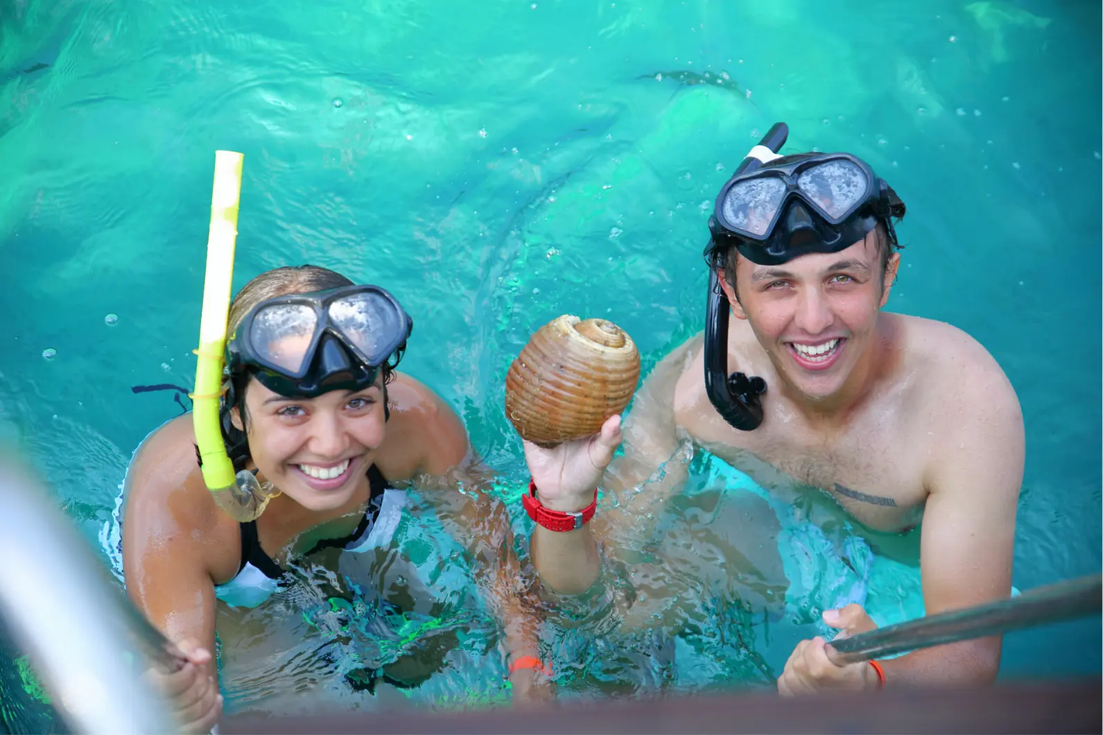 Smiling couple in snorkel masks at the boat ladder, one holding a conch shell