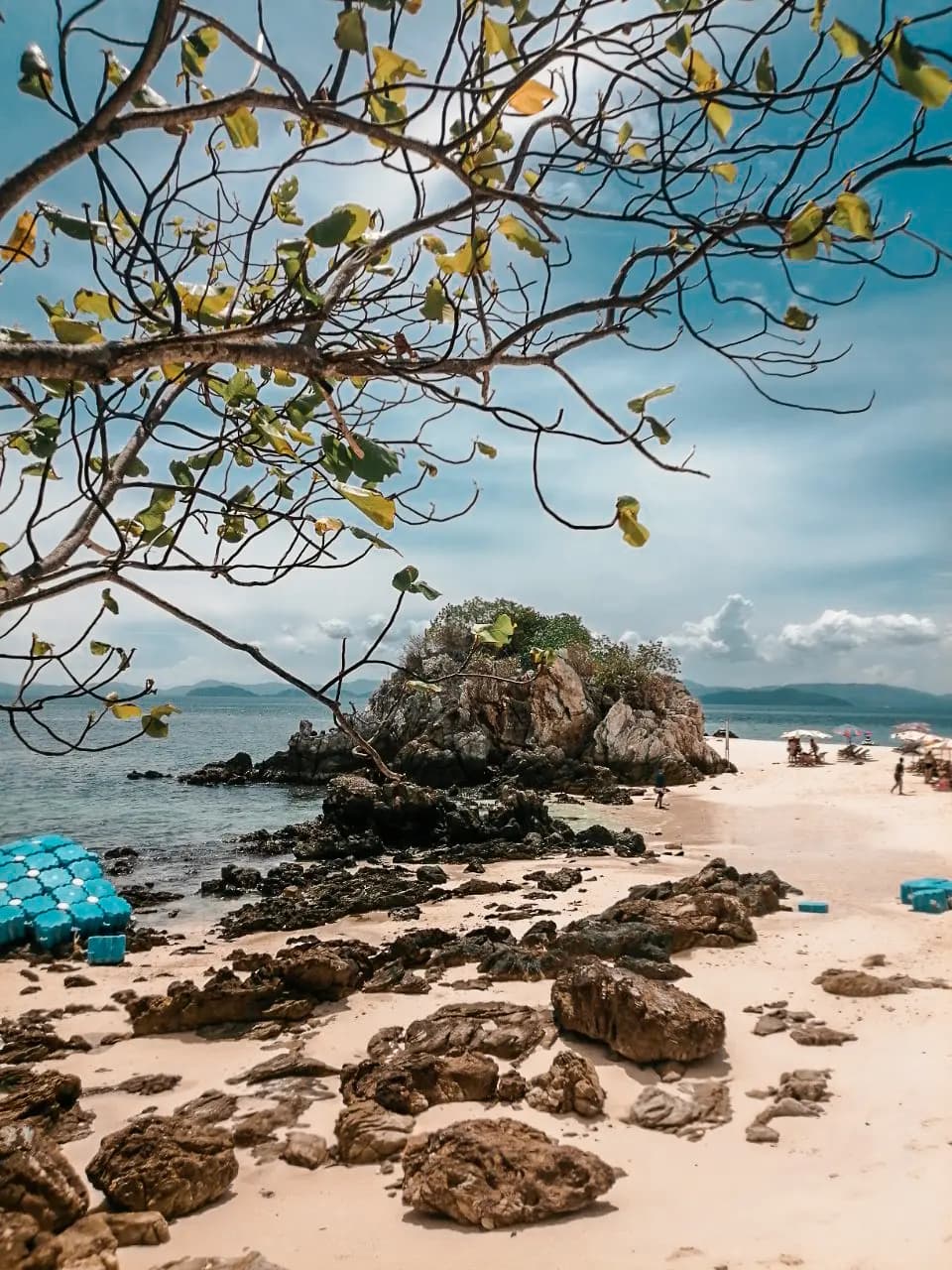 Leafy branches framing a rock-strewn white beach and a shrub-topped boulder outcrop on a Khai island