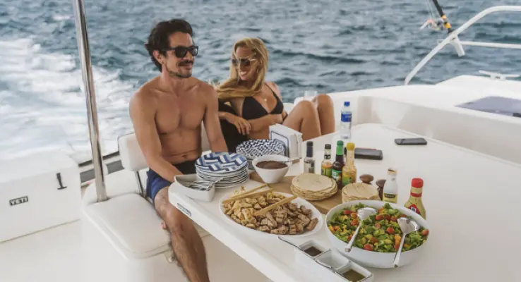 A couple sits at a deck table spread with tortillas, salad and hot sauces aboard the power catamaran