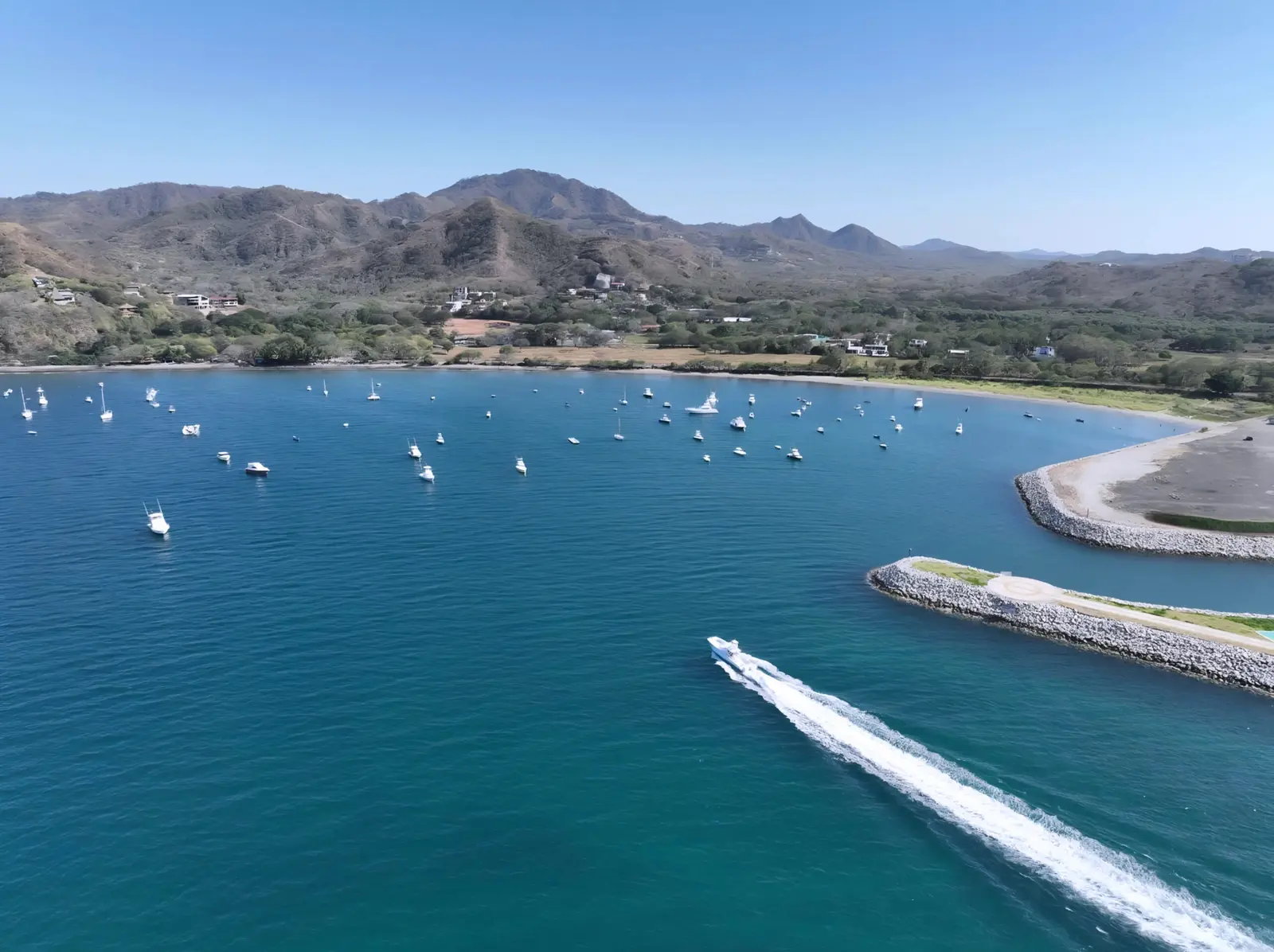 Aerial view of a boat speeding out past the Playa Flamingo breakwater, anchored boats and hills behind