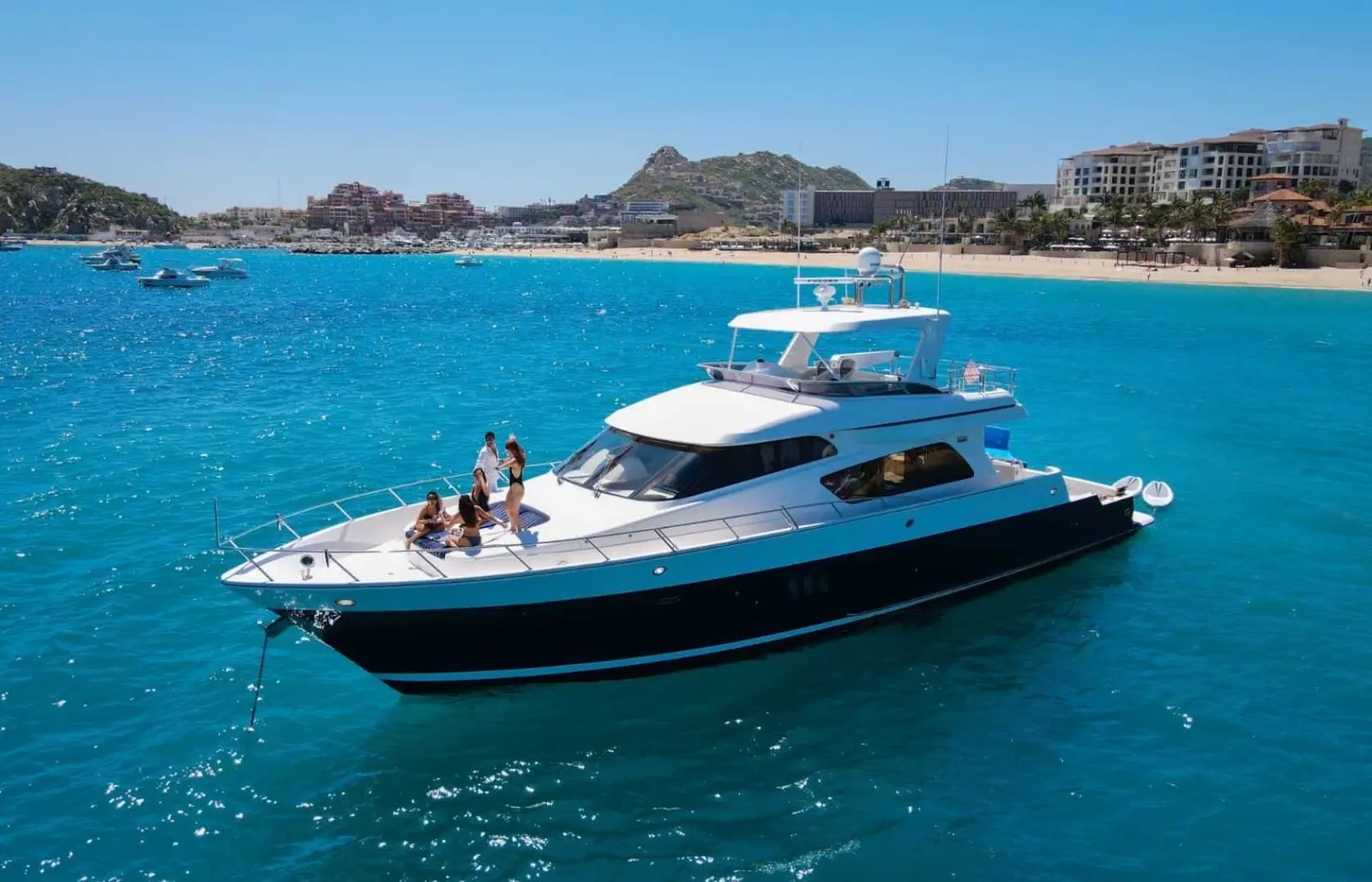 Motor yacht with guests on the bow anchored off a resort-lined beach in Cabo San Lucas