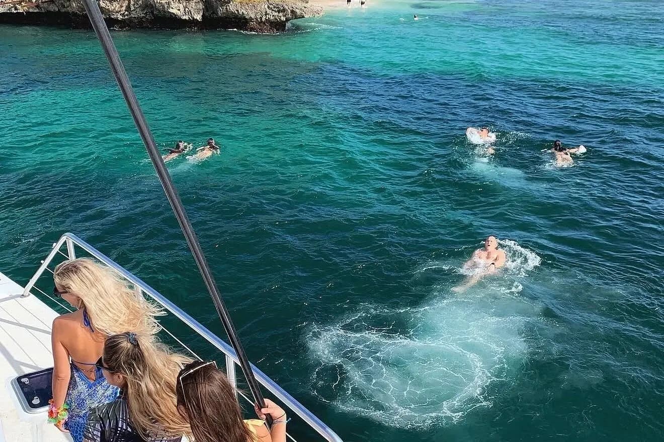 Three women at a catamaran's bow rail watching guests swim in deep green water off a rocky Samaná shore