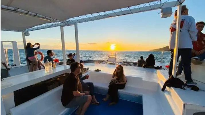 Guests under a catamaran's shade canopy watching the sun set off Los Cabos as one takes a photo