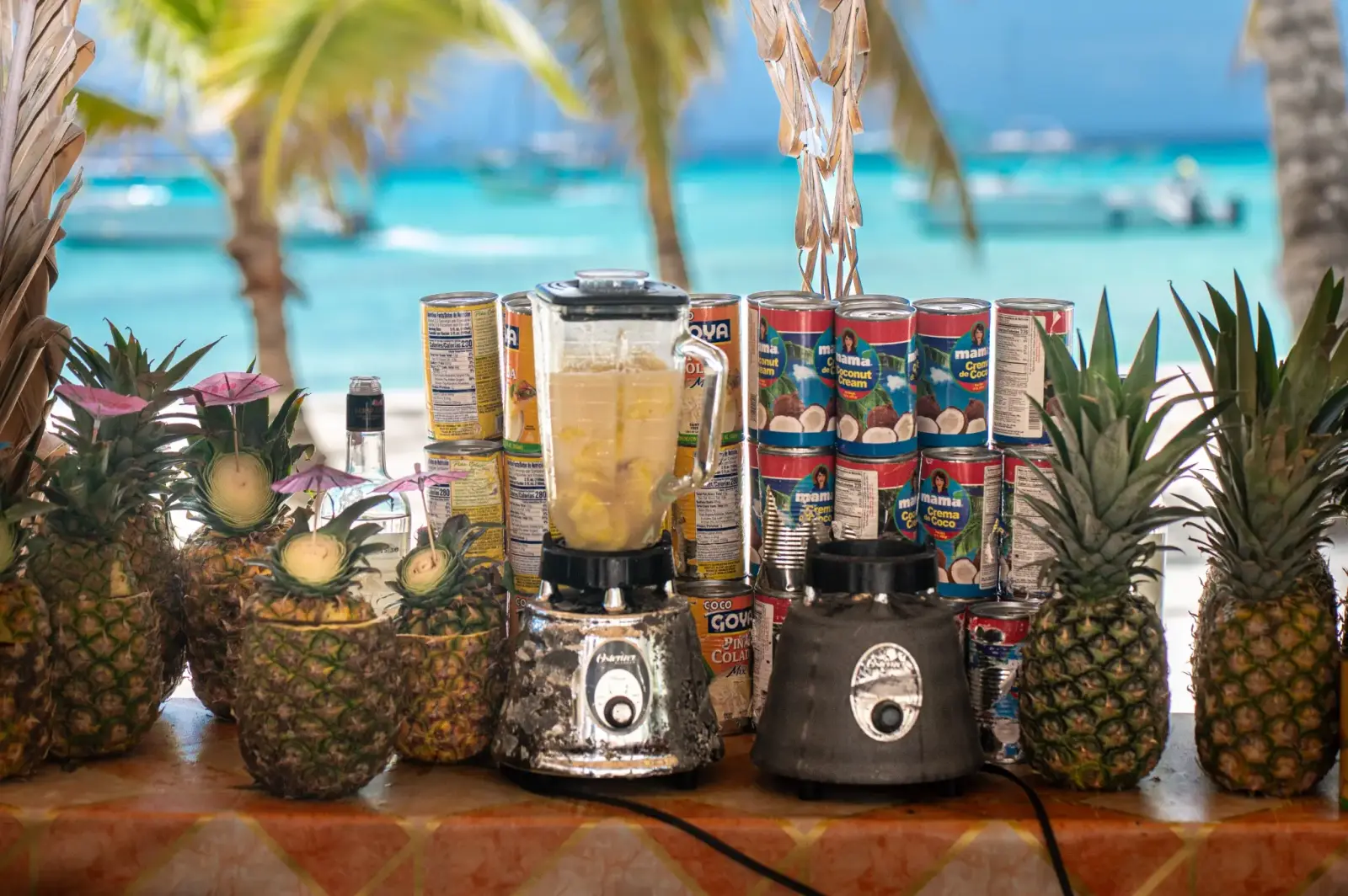 Pineapples, blenders and coconut cream cans set out on a beach bar for blended piña coladas