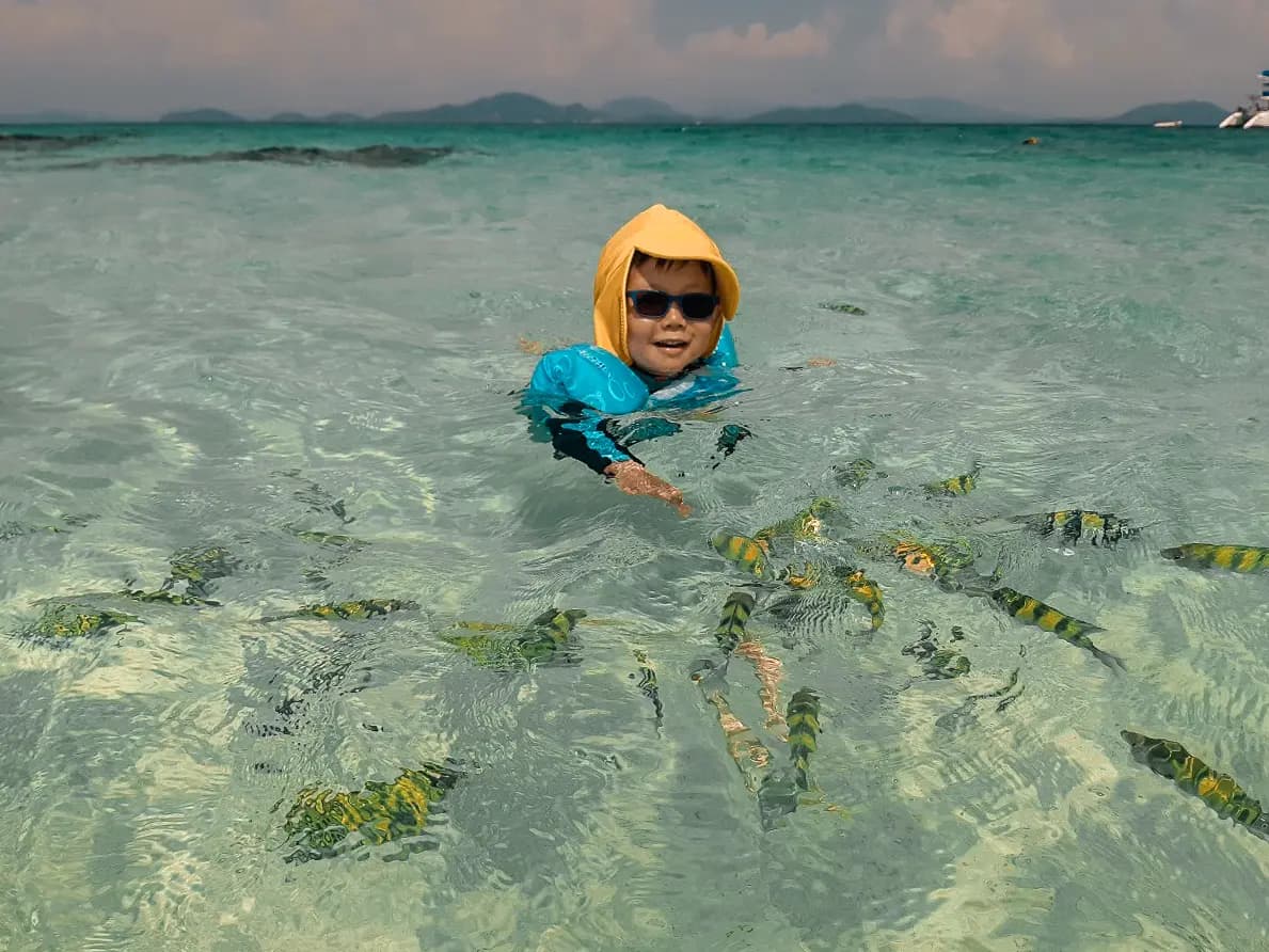 Child in a yellow hood and sunglasses wading among striped fish in the clear shallows off the Khai Islands
