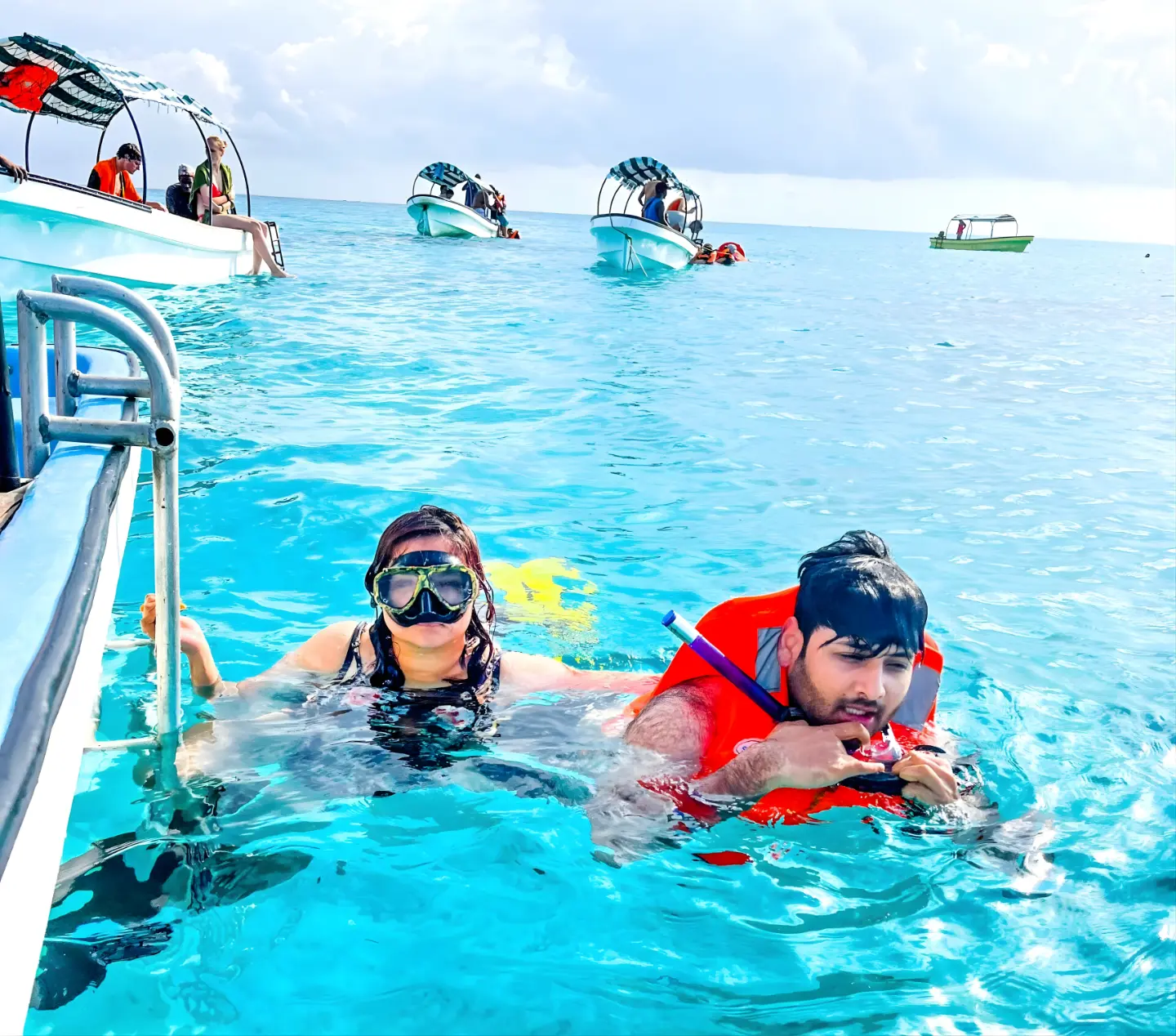 Two snorkelers smiling by a boat's ladder in clear turquoise water, one in an orange life vest