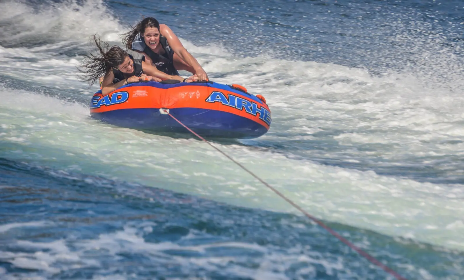 Two riders cling to an inflatable tube bouncing through the boat's foamy wake