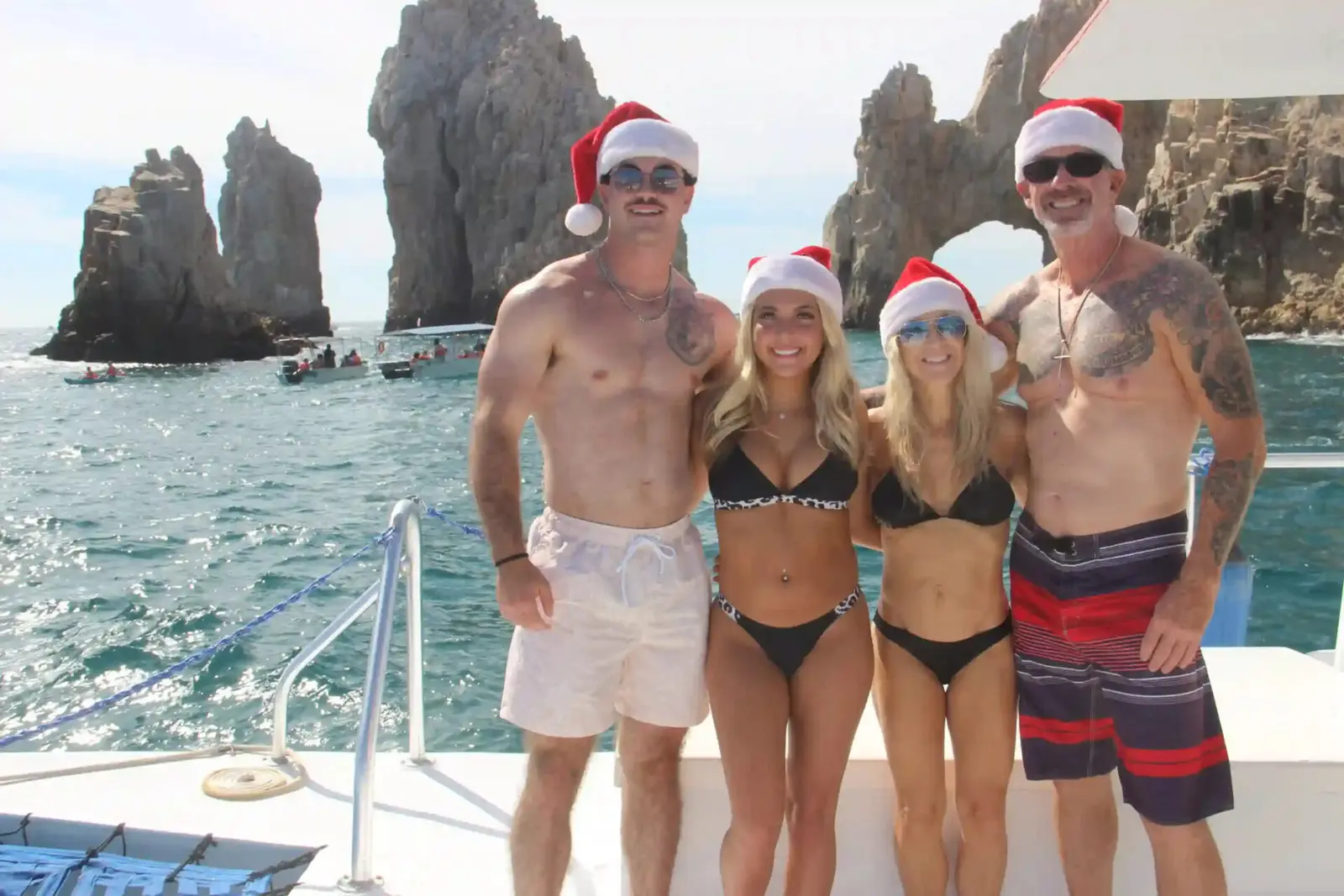 Four guests in Santa hats and swimwear pose on the yacht's deck in front of the Cabo San Lucas arch