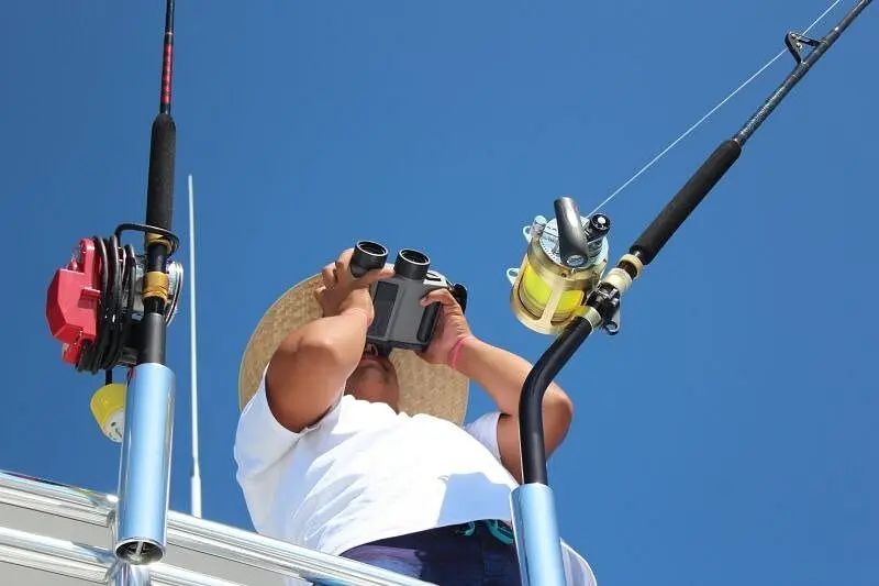 A man in a straw hat scans the water through binoculars beside rigged fishing rods